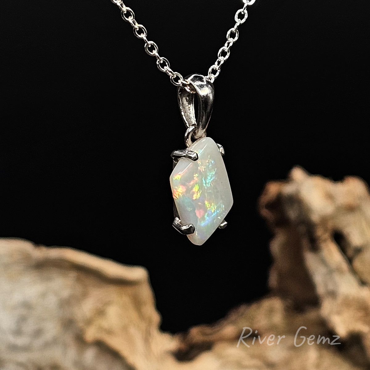 Diamond shaped multi-coloured light opal claw set in silver necklace with large bail against a black background and light drift wood at the base with 'River Gemz' water mark in the bottom right hand corner.