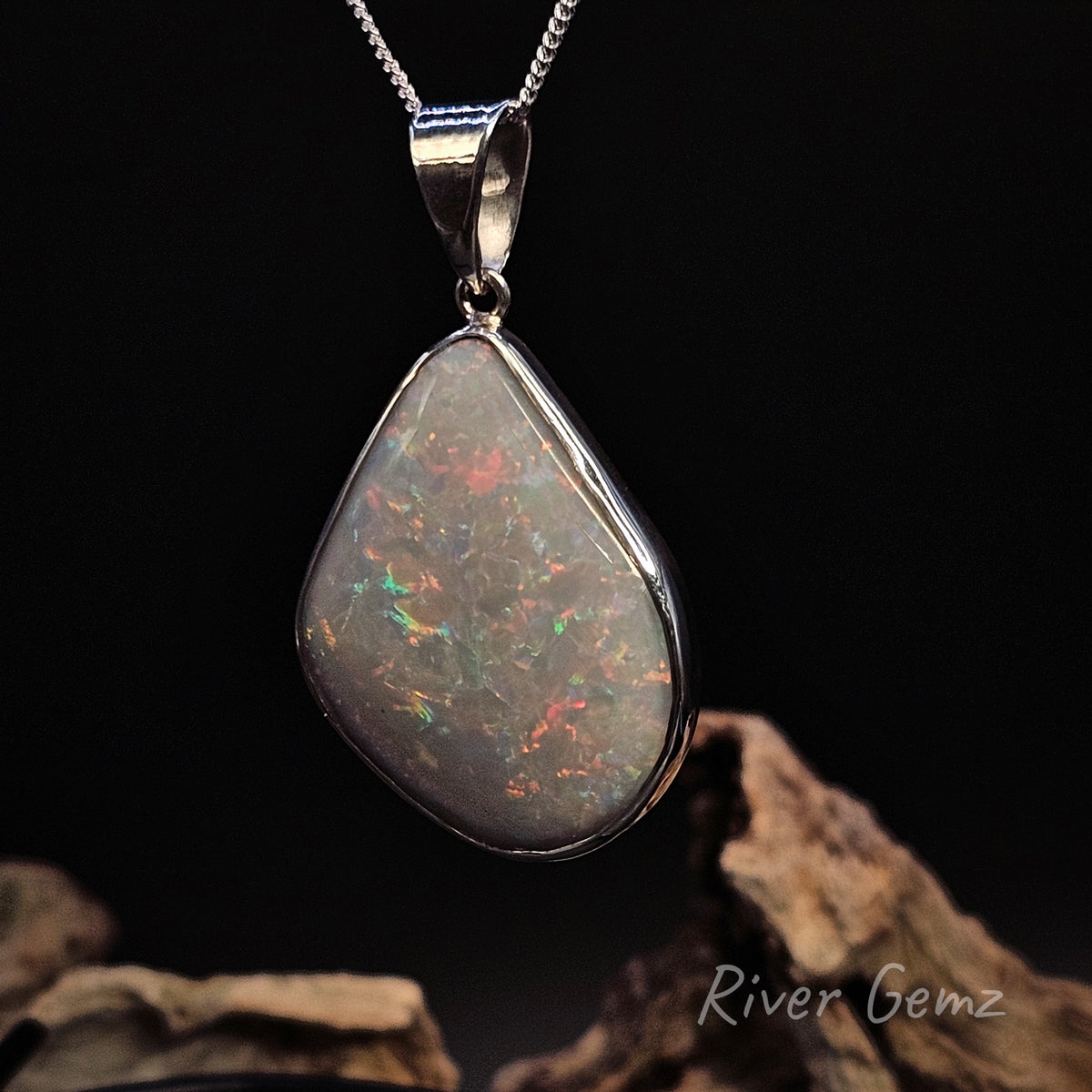 A teardrop-shaped opal pendant in a polished silver bezel setting on a matching silver chain, displayed against a dark background with decorative driftwood at the bottom. The opal displays a play-of-color with flashes of orange, green, and blue on a dark base.