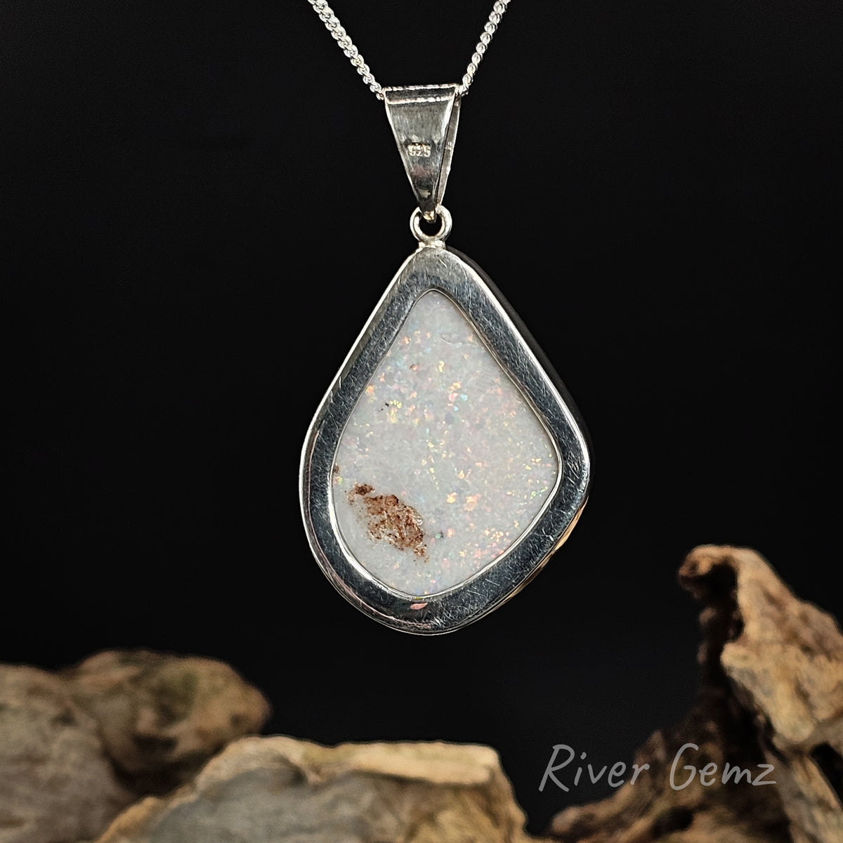 Rear view of pendant shows light base tone of the white opal set in it. The pendant is displayed against a dark background with decorative driftwood at the bottom, and the 'River Gemz' brand logo is visible in the lower right corner.