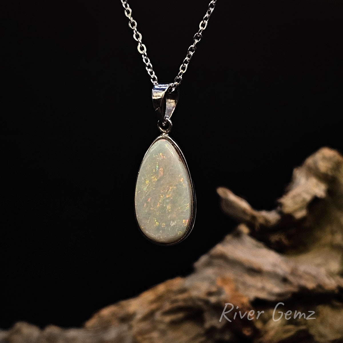 A silver pendant necklace featuring a teardrop-shaped opal gemstone with iridescent internal flashes, displayed against a black background with a piece of driftwood and a 'River Gemz' watermark.