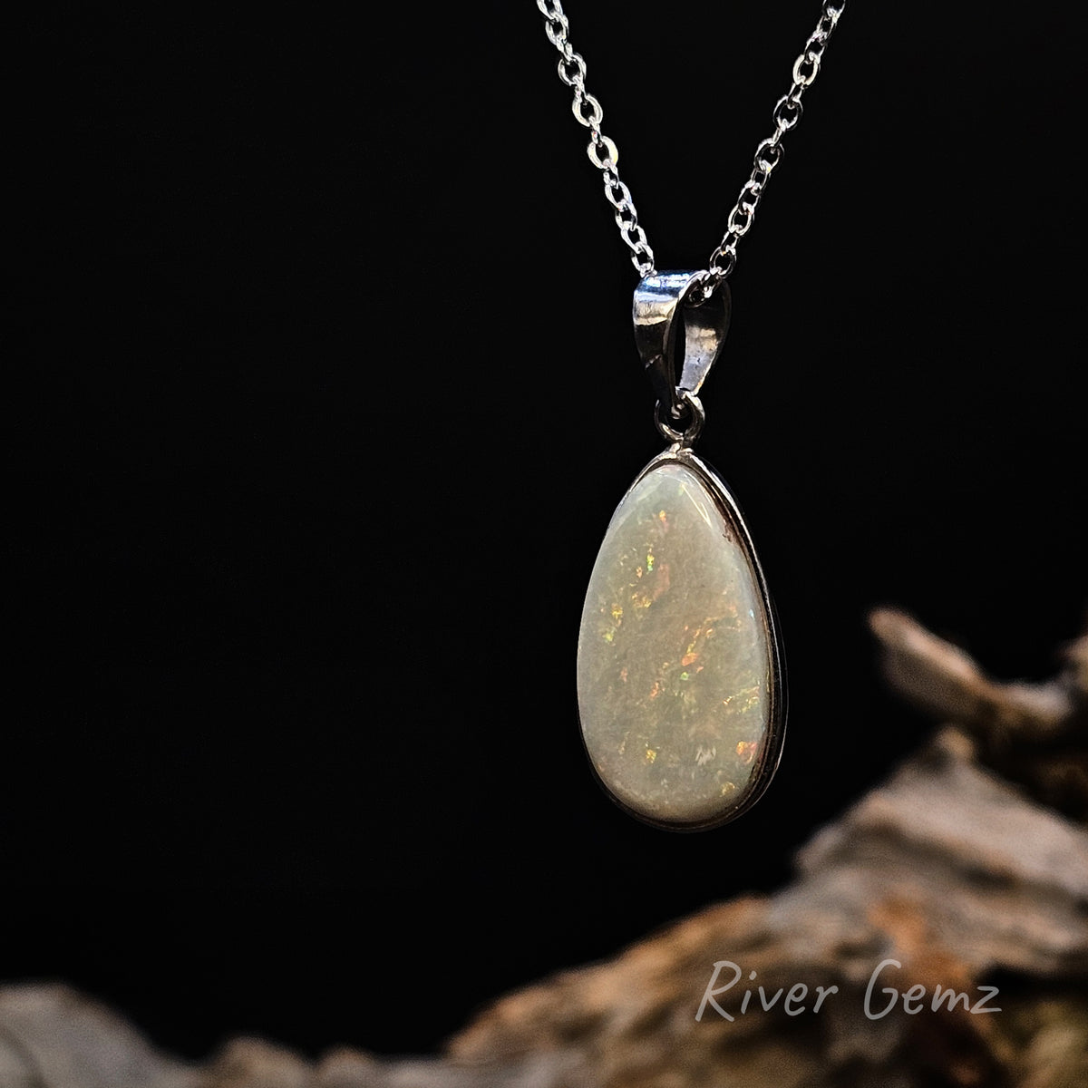 A silver teardrop pendant necklace featuring a milky white opal with vibrant flashes of orange, green, and blue, hanging against a solid black background with a piece of driftwood at the bottom.