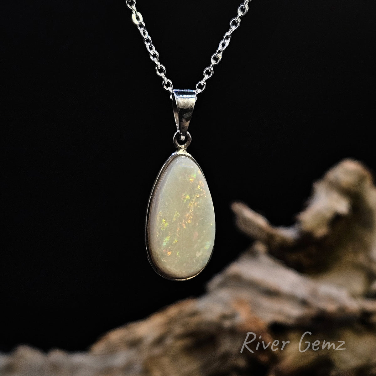 A silver teardrop pendant necklace featuring a white opal gemstone with iridescent flashes, hanging from a silver chain against a black background with a piece of driftwood at the bottom right. The watermark 'River Gemz' is visible in the lower right corner.