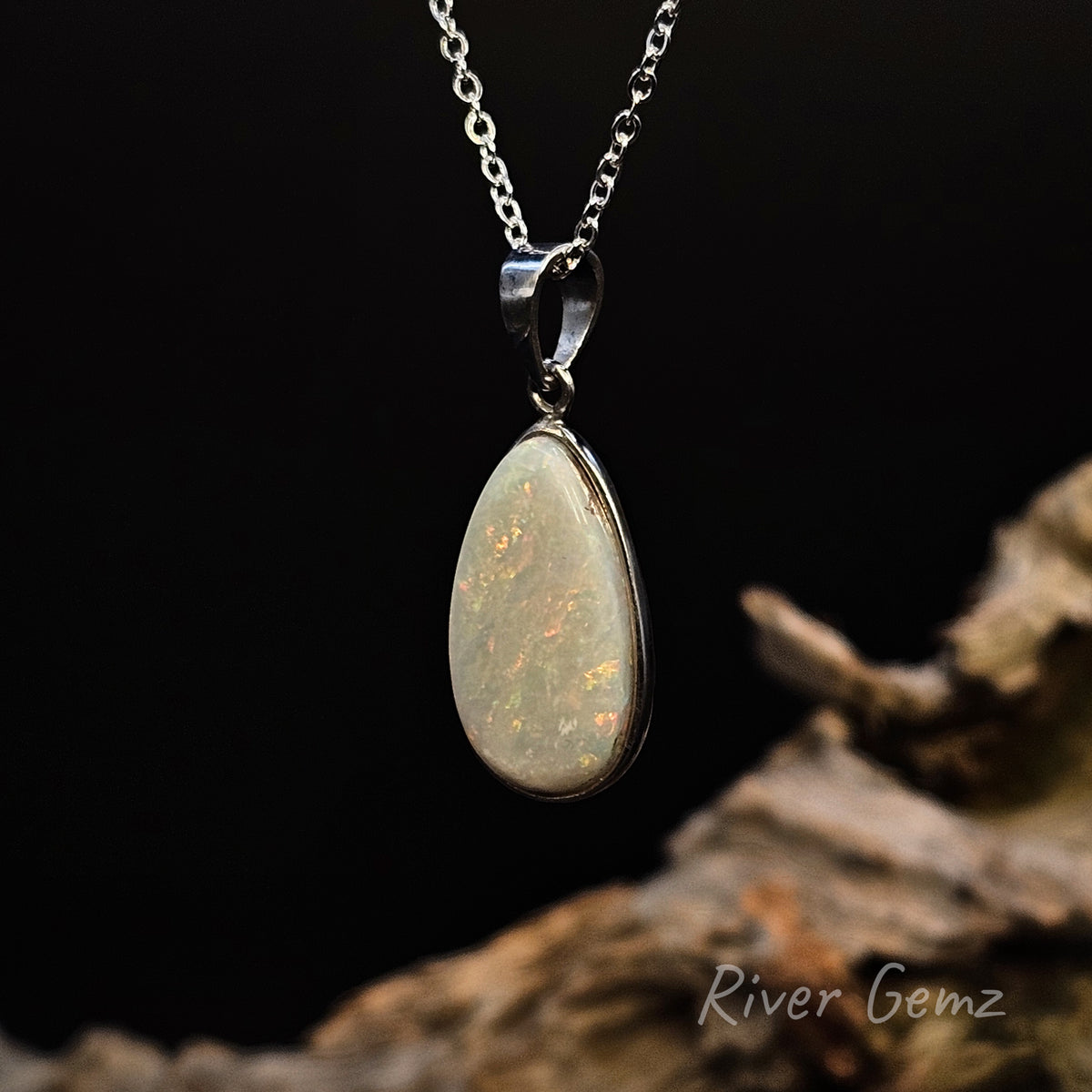 A silver pendant necklace featuring a teardrop-shaped opal gemstone with natural play-of-color, displayed against a dark background with a piece of driftwood.