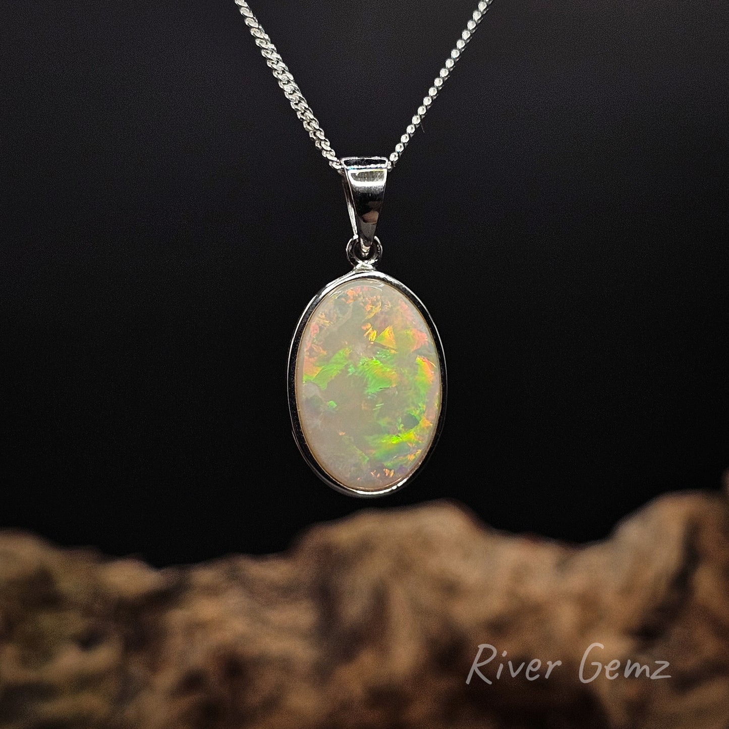 Oval shaped multi-coloured light opal besel set in classic style silver pendant against a black background and blurred timber base with 'River Gemz' logo.