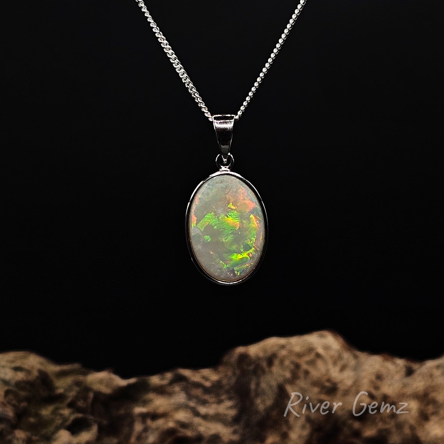 Large oval shaped multi-colour light opal besel set in sterling silver pendant against black background and drift wood base with 'River Gemz' watermark.