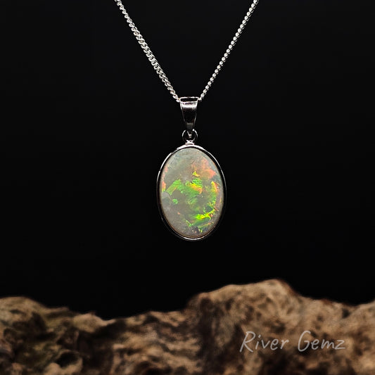 Large oval shaped multi-colour light opal besel set in sterling silver pendant against black background and drift wood base with 'River Gemz' watermark.
