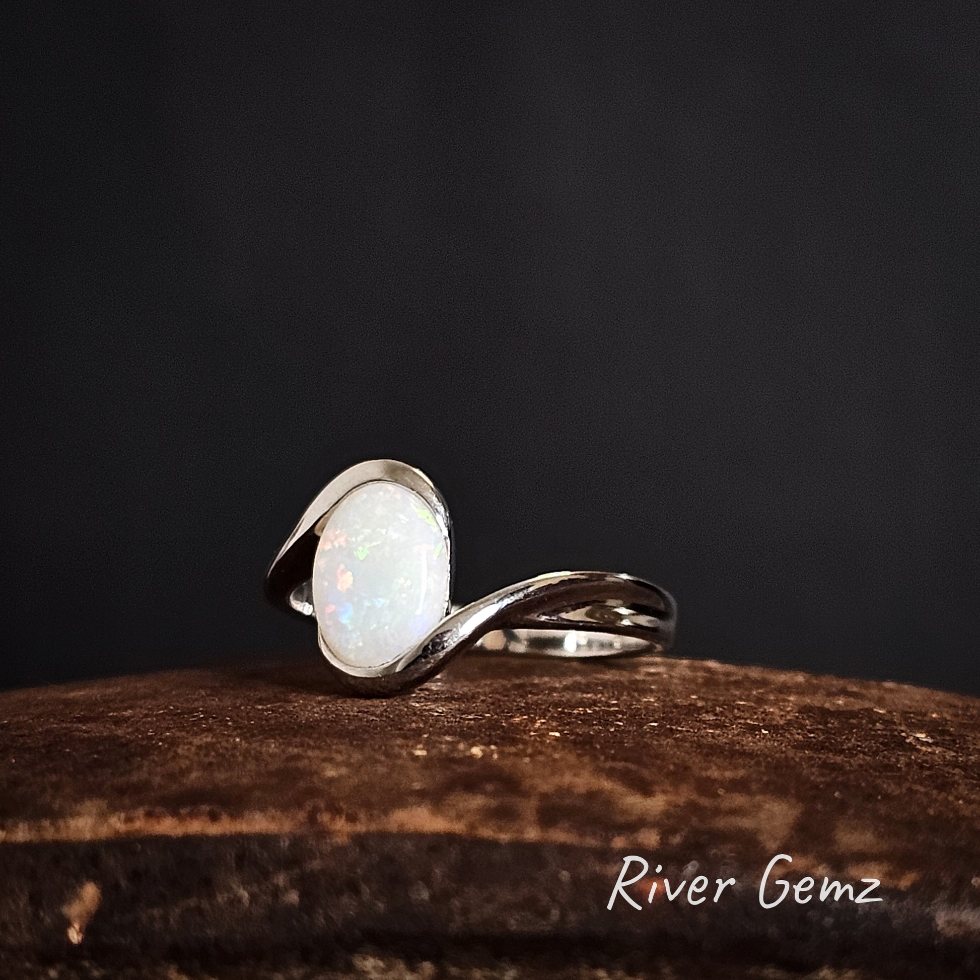 Silver ring with a partial besel set white opal stone on a dark background, branded 'River Gemz'.