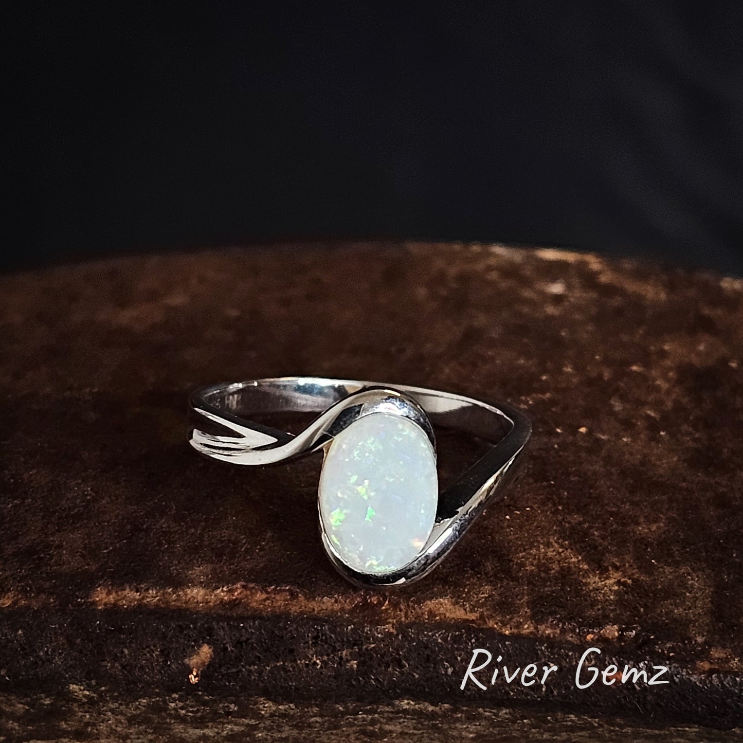 Silver ring with a light multi-coloured opal stone on a dark brown surface, branded 'River Gemz'.