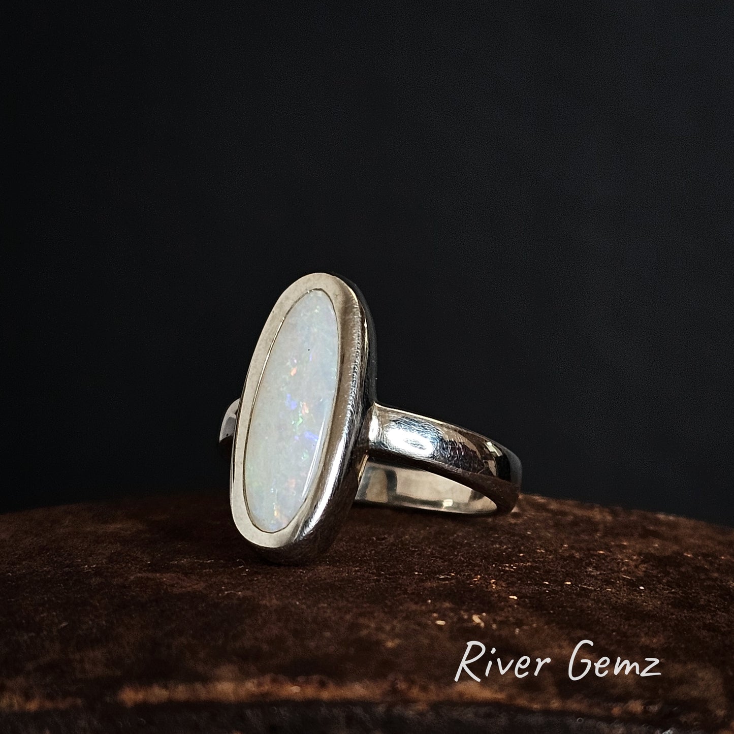 Silver ring with an oval shaped white opal on a dark background, branded 'River Gemz'.