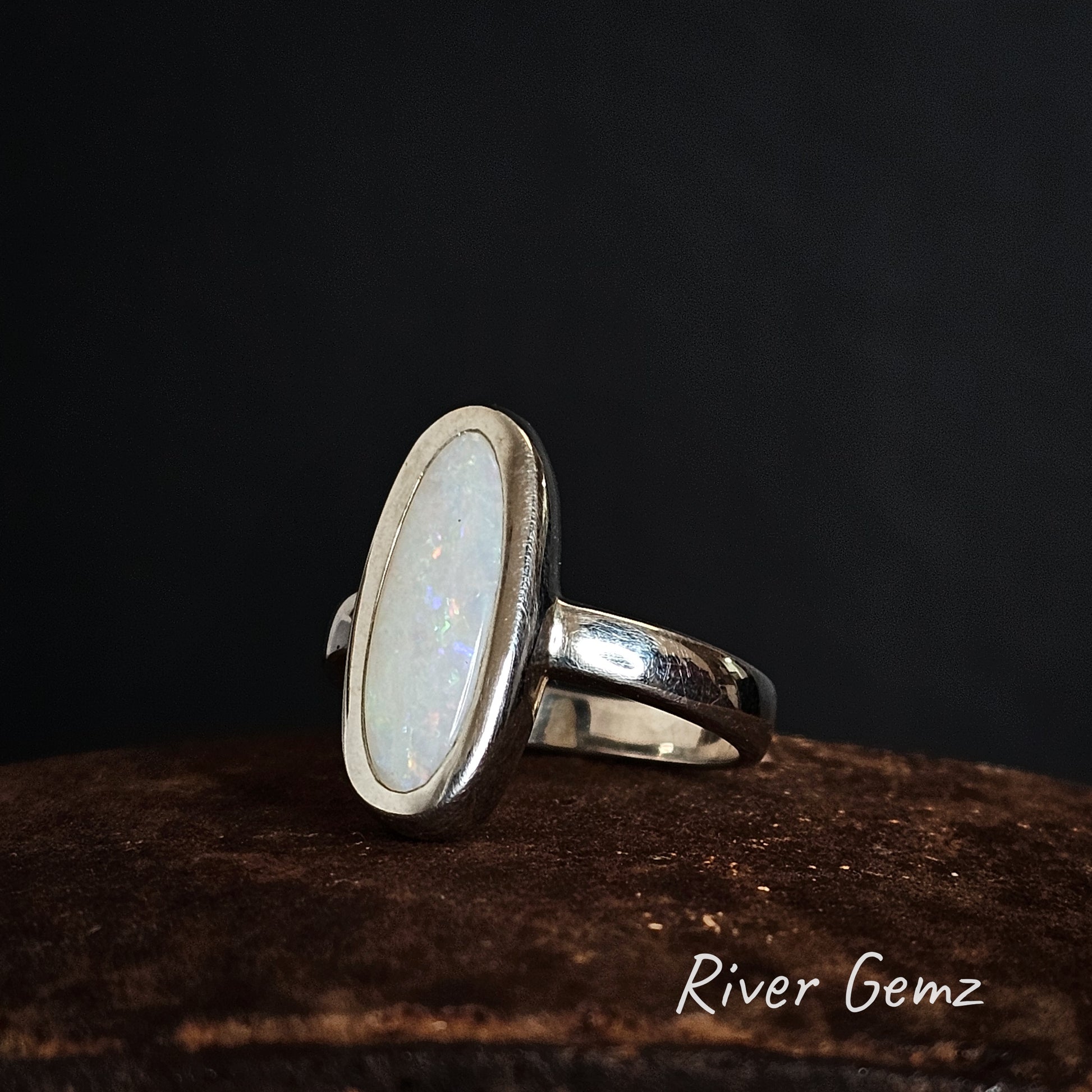 Silver ring with an oval shaped white opal on a dark background, branded 'River Gemz'.