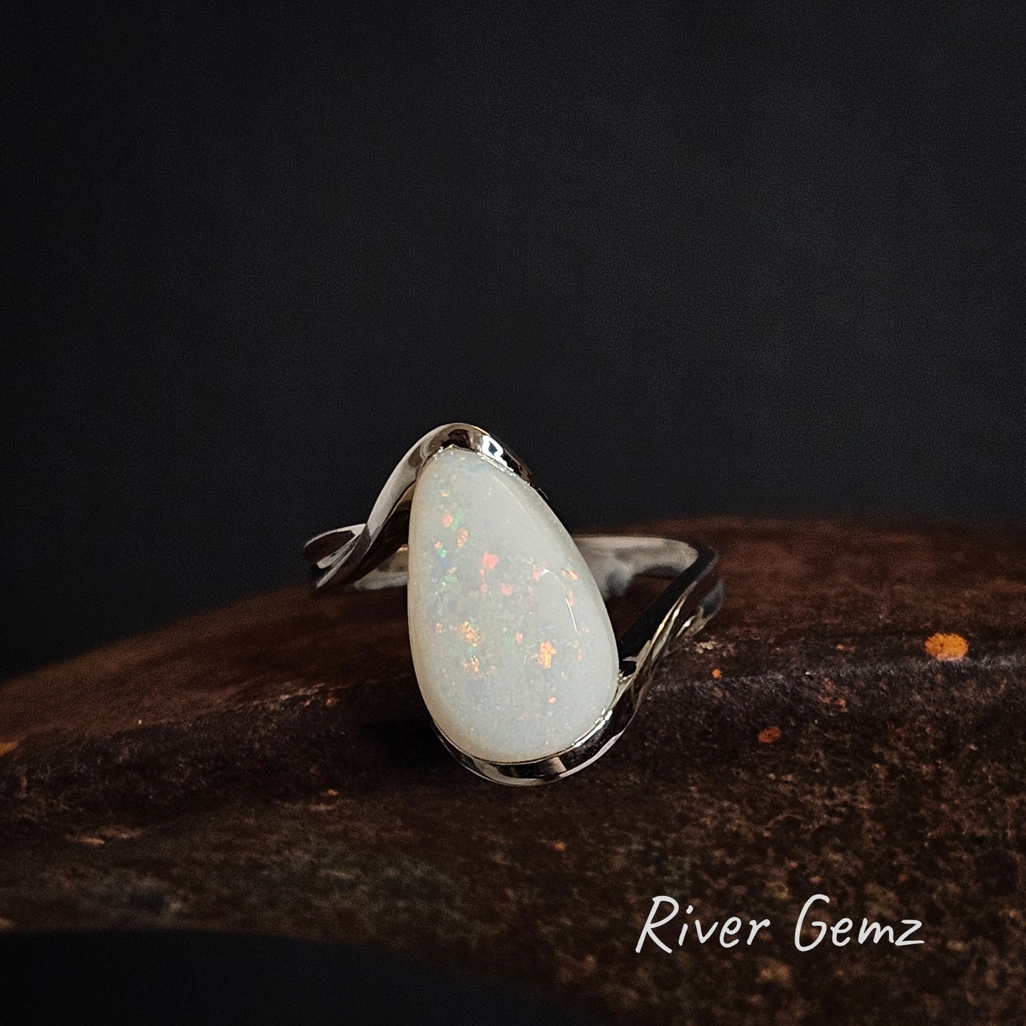 Silver ring with a teardrop shaped light opal stone on a dark surface, branded 'River Gemz'.