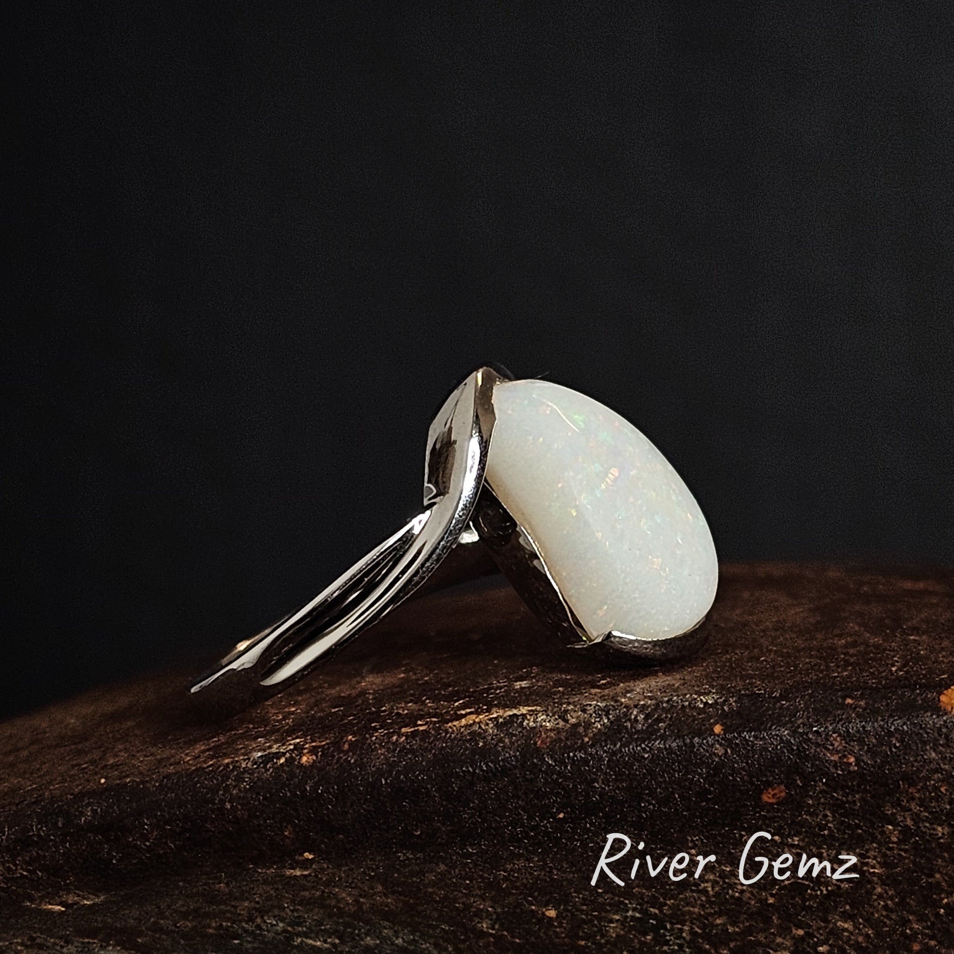 Silver ring with a high cabochon cut white opal stone on a dark background, branded 'River Gemz'.
