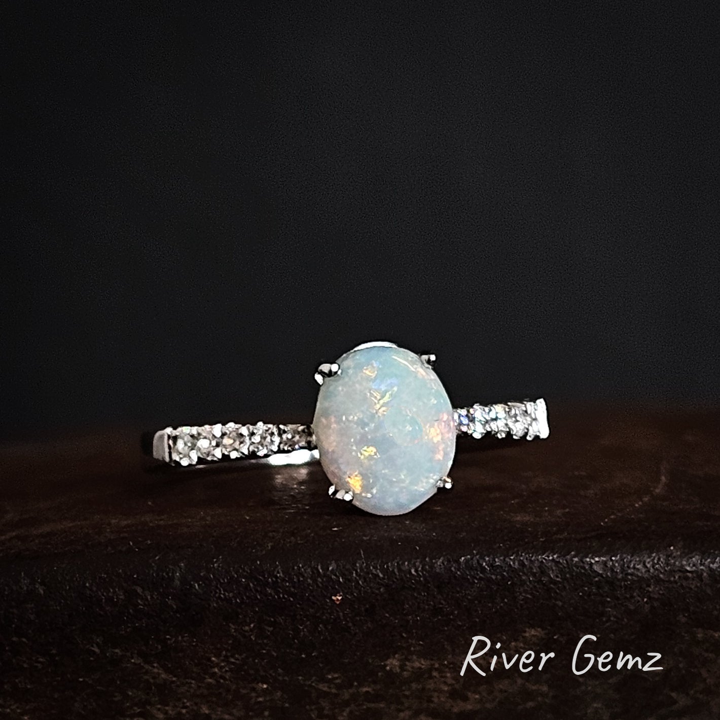 Opal ring with white topaz accents on a dark surface, branded 'River Gemz'.