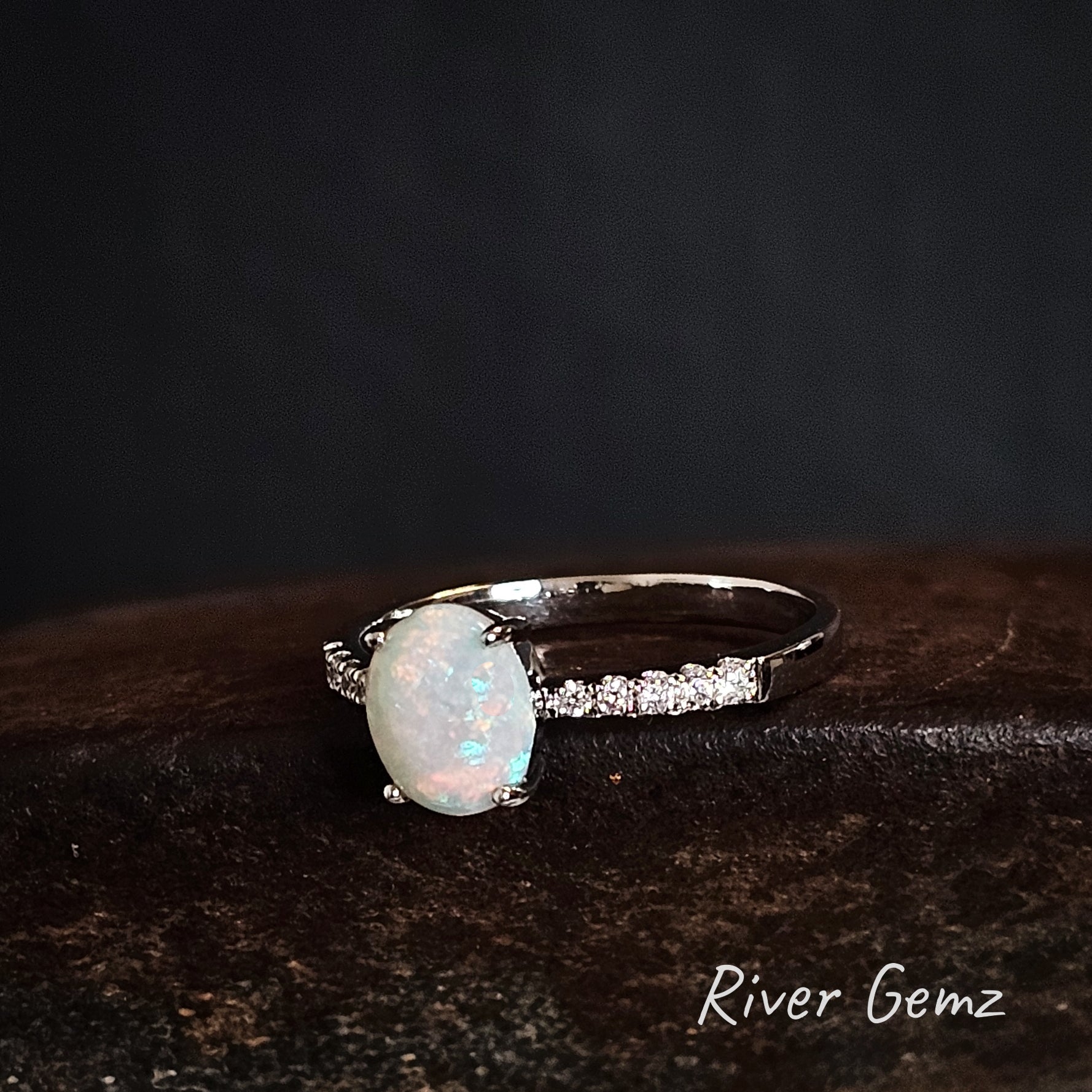 Oval shaped white opal and topaz stones in a silver ring on a dark background, branded 'River Gemz'.