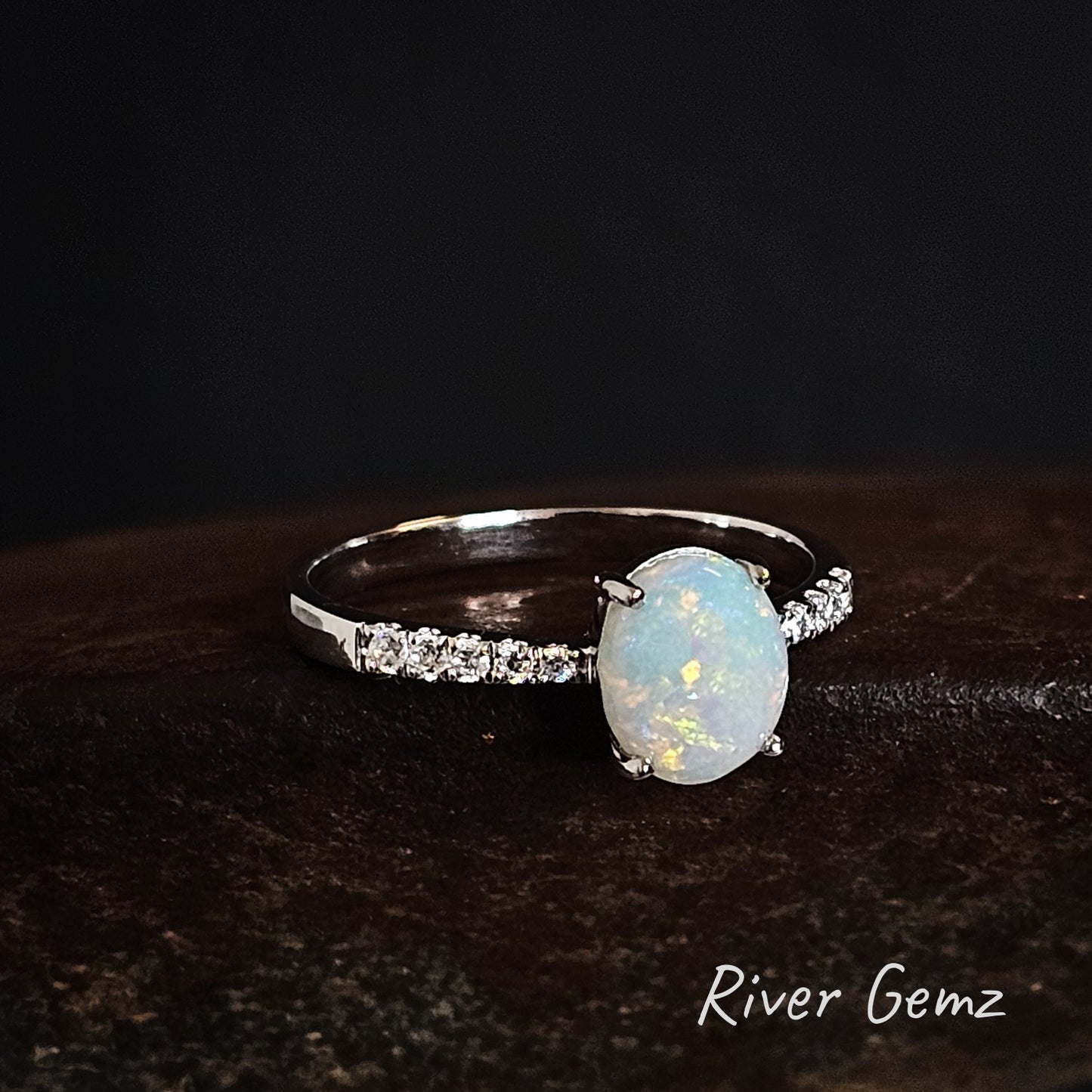 Silver ring with an multi-coloured white opal stone on a dark background, branded 'River Gemz'.