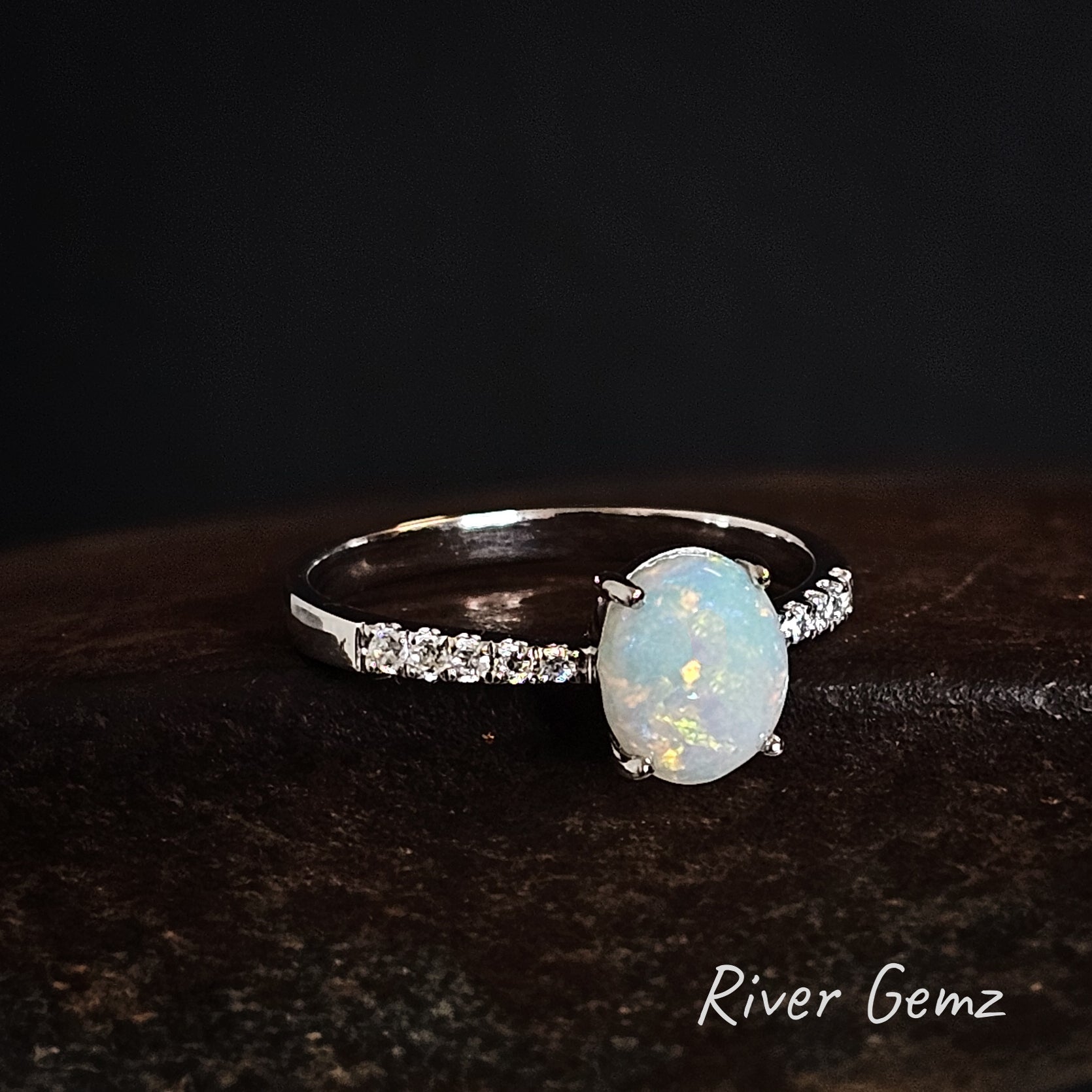 Silver ring with an multi-coloured white opal stone on a dark background, branded 'River Gemz'.