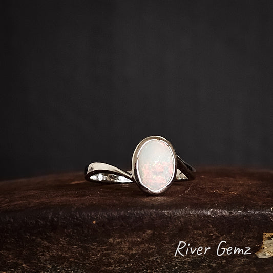 Oval shaped white opal besel set in sterling silver ring on a dark surface branded 'River Gemz'.