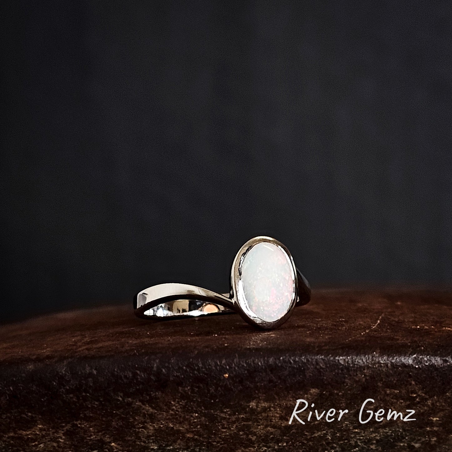 Oval shaped light opal set in sterling silver ring on a rusty brown surface with 'River Gemz' branding.