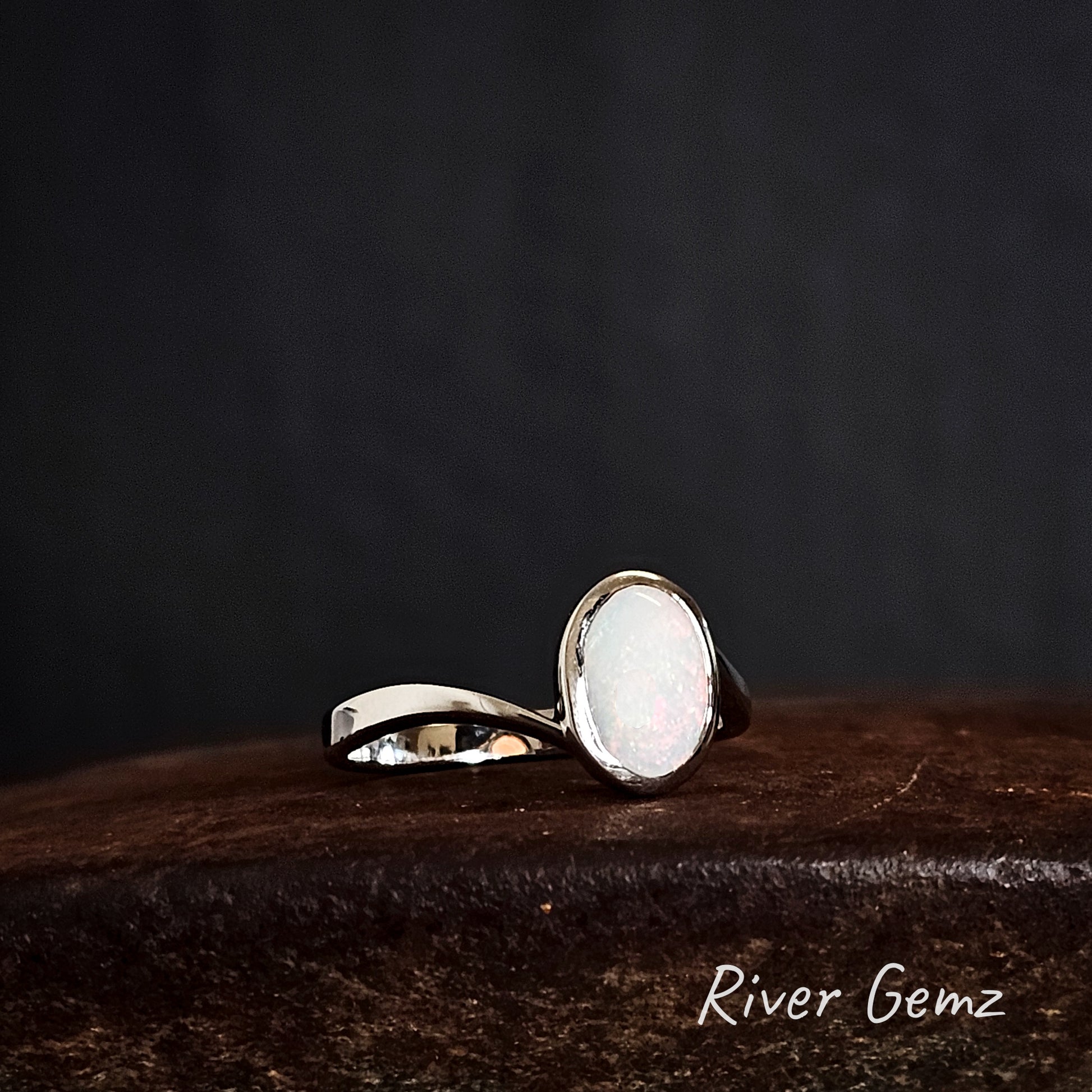 Oval shaped light opal set in sterling silver ring on a rusty brown surface with 'River Gemz' branding.