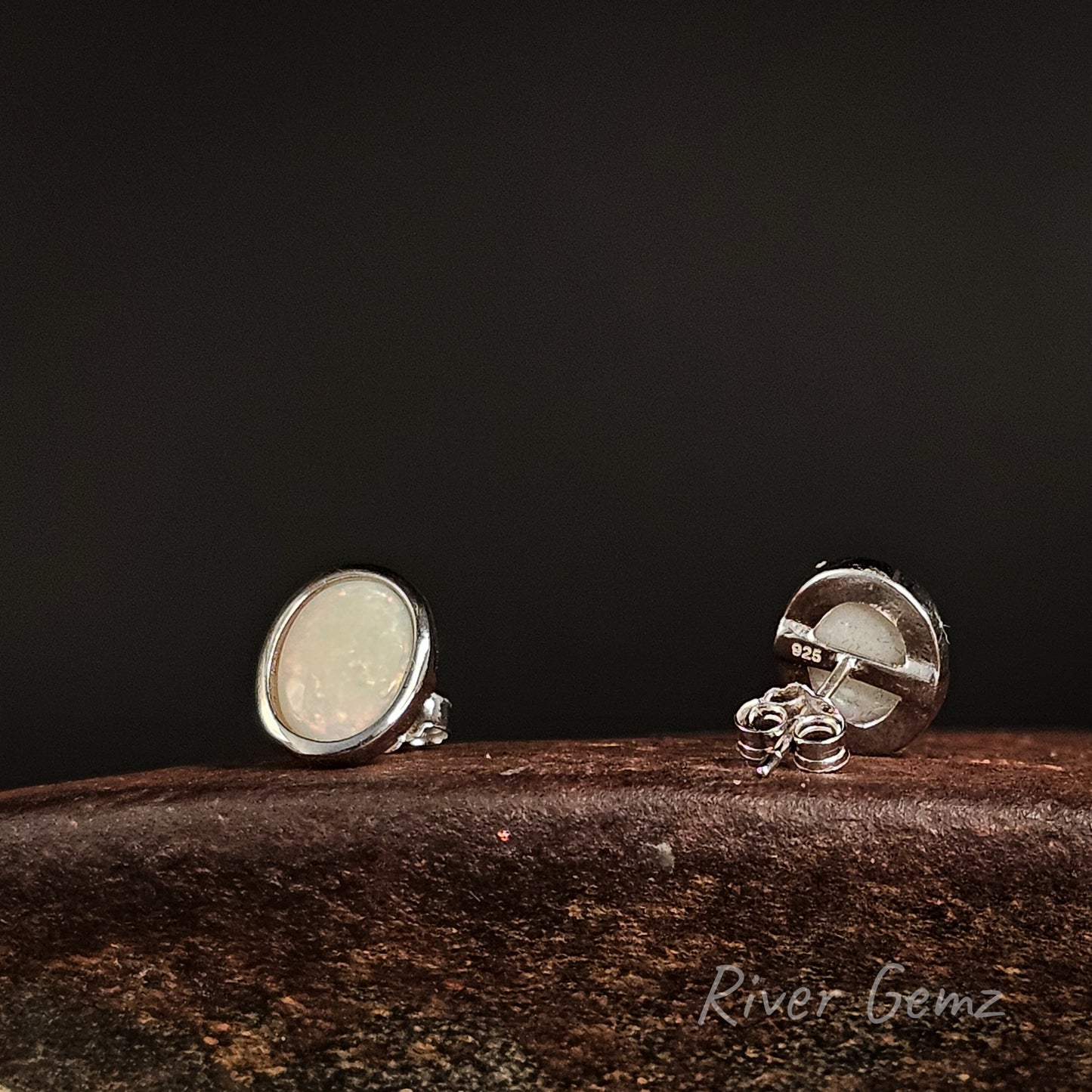 Pair of silver earrings on a dark surface with 'River Gemz' branding.