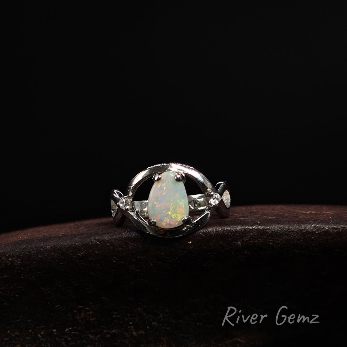 A silver ring featuring a teardrop-shaped iridescent opal gemstone, displayed on a dark brown textured surface against a solid black background. The brand name 'River Gemz' is visible in the bottom right corner.
