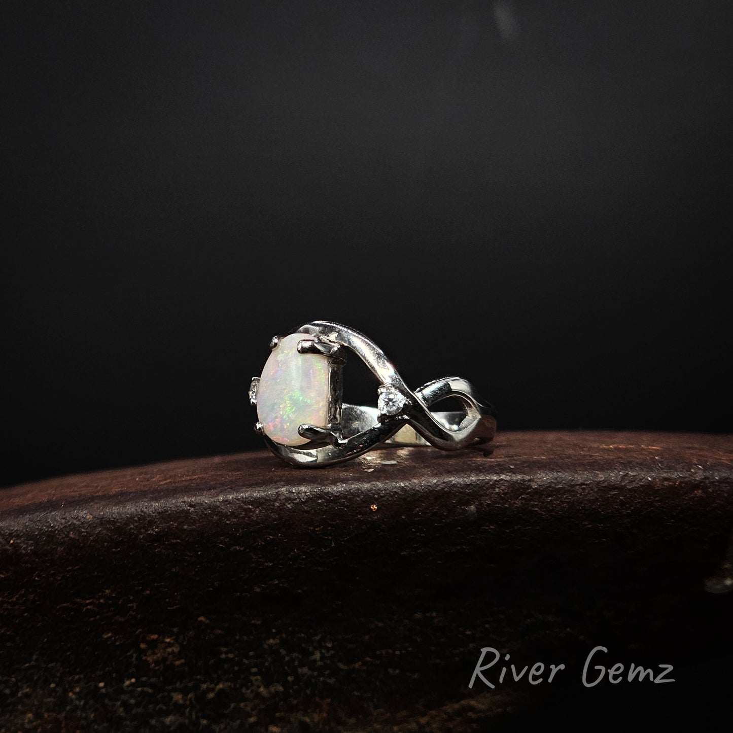 A sterling silver ring featuring a large oval cabochon opal with iridescent white and green flashes, set in a polished infinity-style band. The ring is displayed on a dark, textured wooden surface against a dark background, with the brand name 'River Gemz' visible in the bottom right corner.