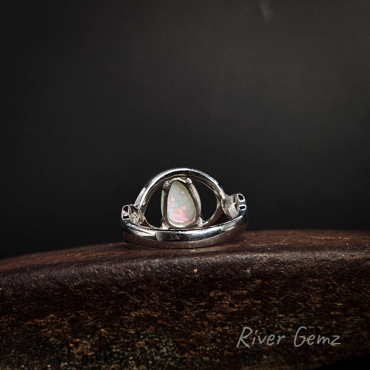 Rear view of silver ring showing the light base tone of the white opal, resting on a dark textured wooden surface against a dark background. The brand name 'River Gemz' is visible in the bottom right corner.