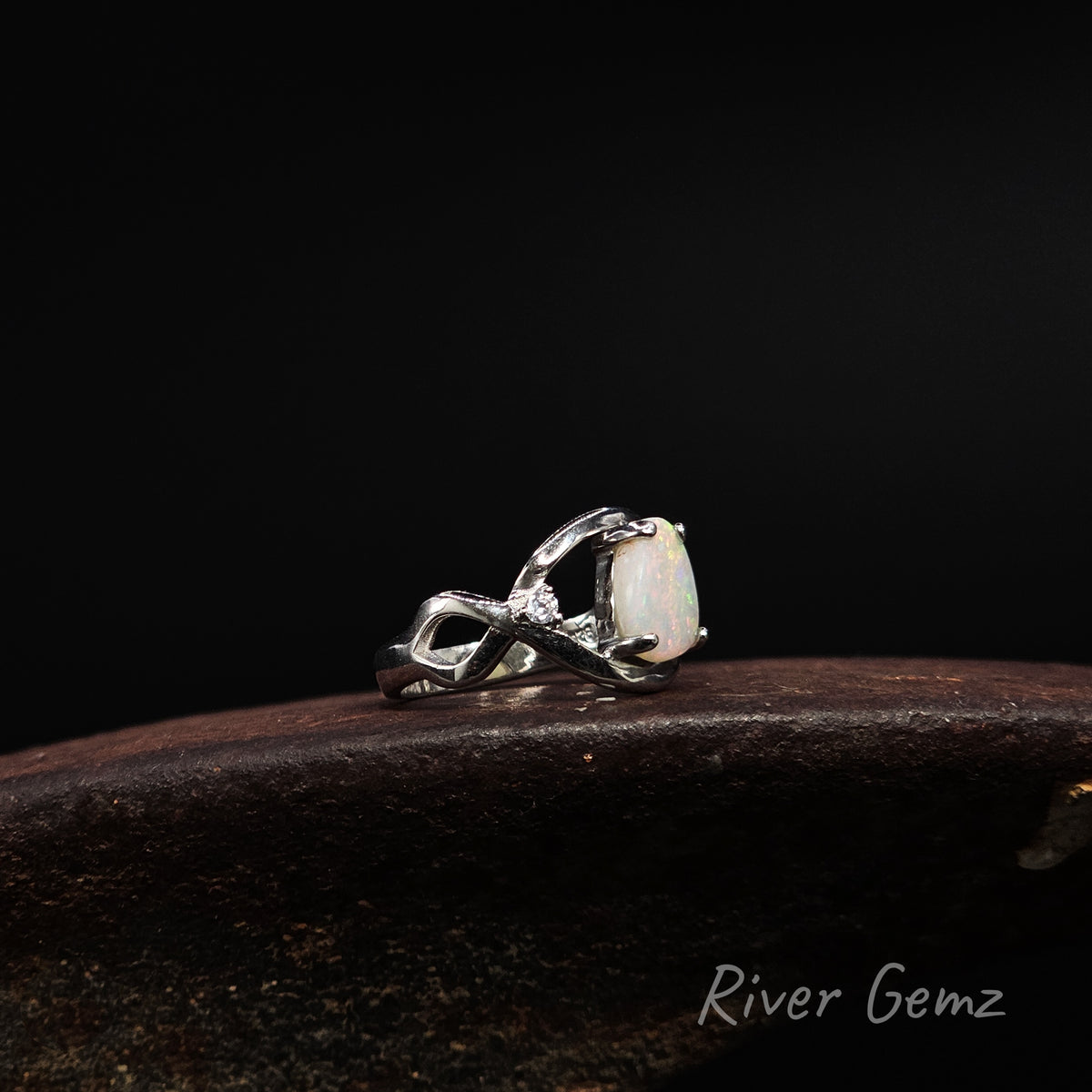 A sterling silver ring featuring a claw-set white opal and a small clear accent stone, resting on a dark wooden surface against a black background. The 'River Gemz' brand name is visible in the bottom right corner.