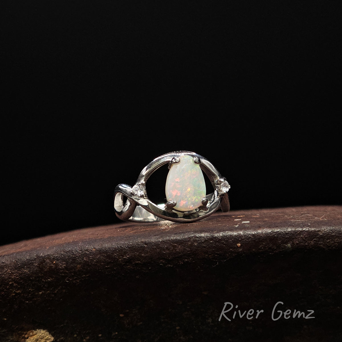 A silver ring featuring a teardrop-shaped white opal with iridescent flashes, displayed on a dark wooden surface against a black background. The brand name 'River Gemz' is visible in the bottom right corner.