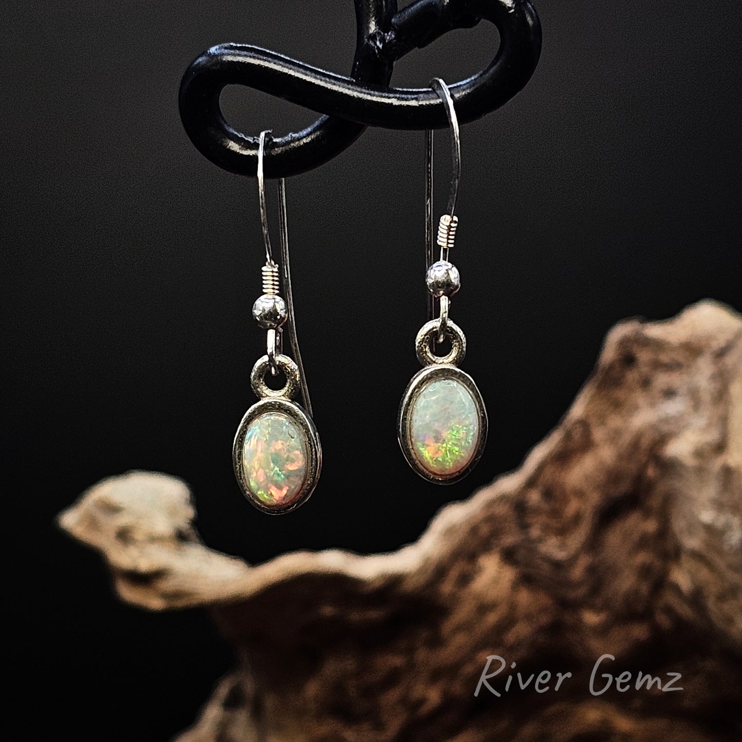 Multi-coloured light opals set in silver hook earrings on dark background with River Gemz branding.
