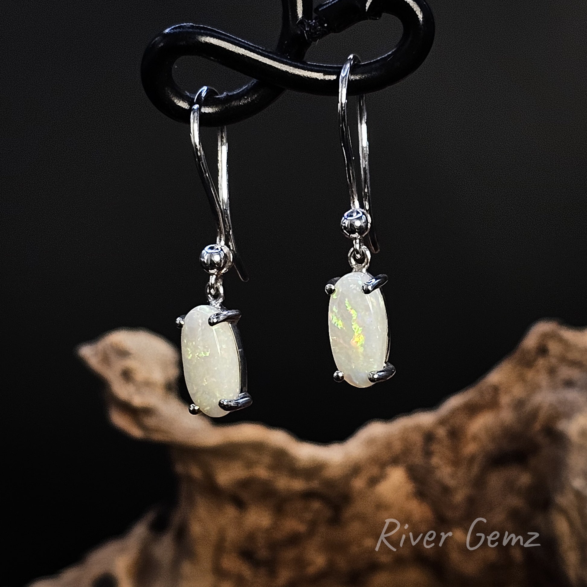 Oval shaped light opal in silver French hooks on a dark background with 'River Gemz' branding.