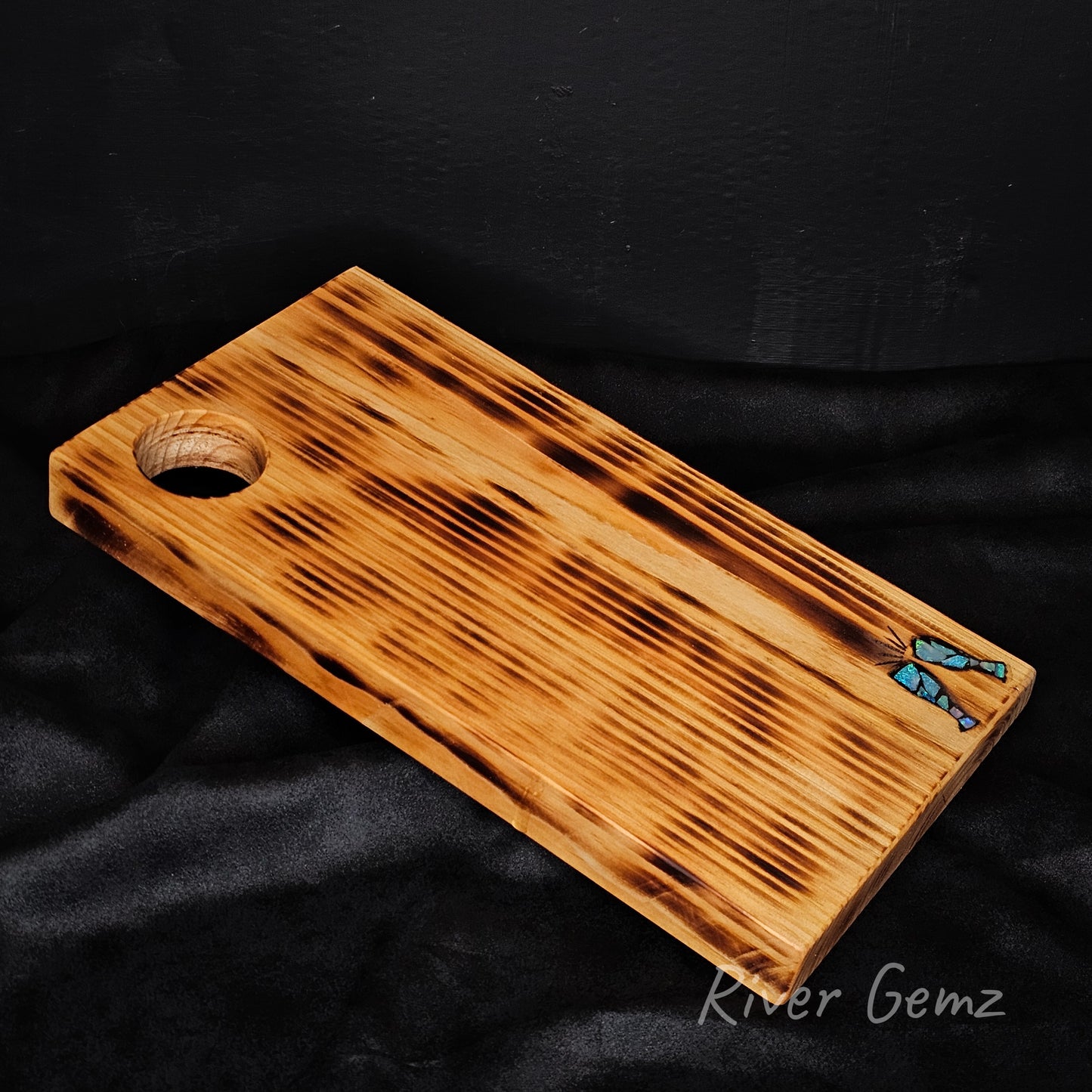 Wooden cheese board with champagne glasses opal inlayed design on a black background, branded 'River Gemz'.