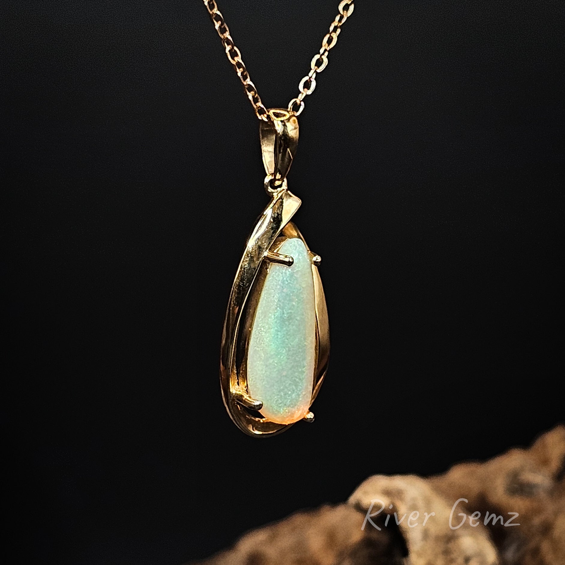 Gold pendant with a teardrop-shaped gemstone on a dark background, featuring 'River Gemz' branding.