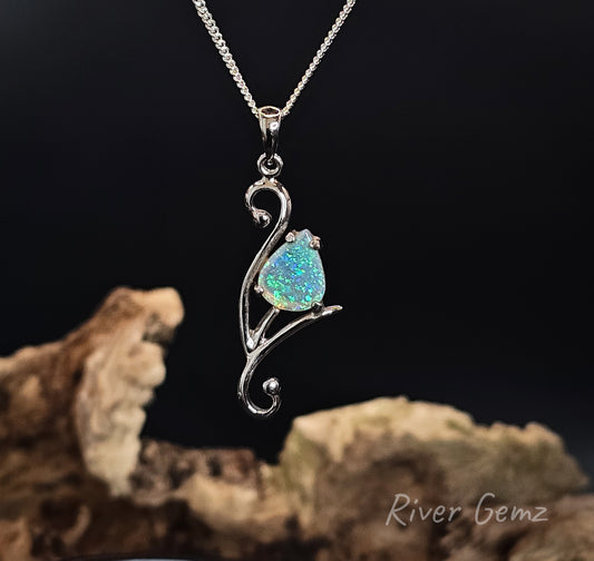 A silver pendant necklace featuring a teardrop-shaped iridescent opal gemstone set in a stylized branch-like silver frame, hanging from a silver chain against a dark background with driftwood props. The watermark 'River Gemz' is visible in the bottom right corner.