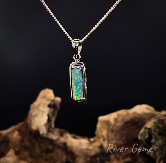 A silver rectangular pendant necklace featuring a vibrant, iridescent opal gemstone, displayed on a dark background with a piece of driftwood in the foreground. The brand name 'River Gemz' is visible in the bottom right corner.
