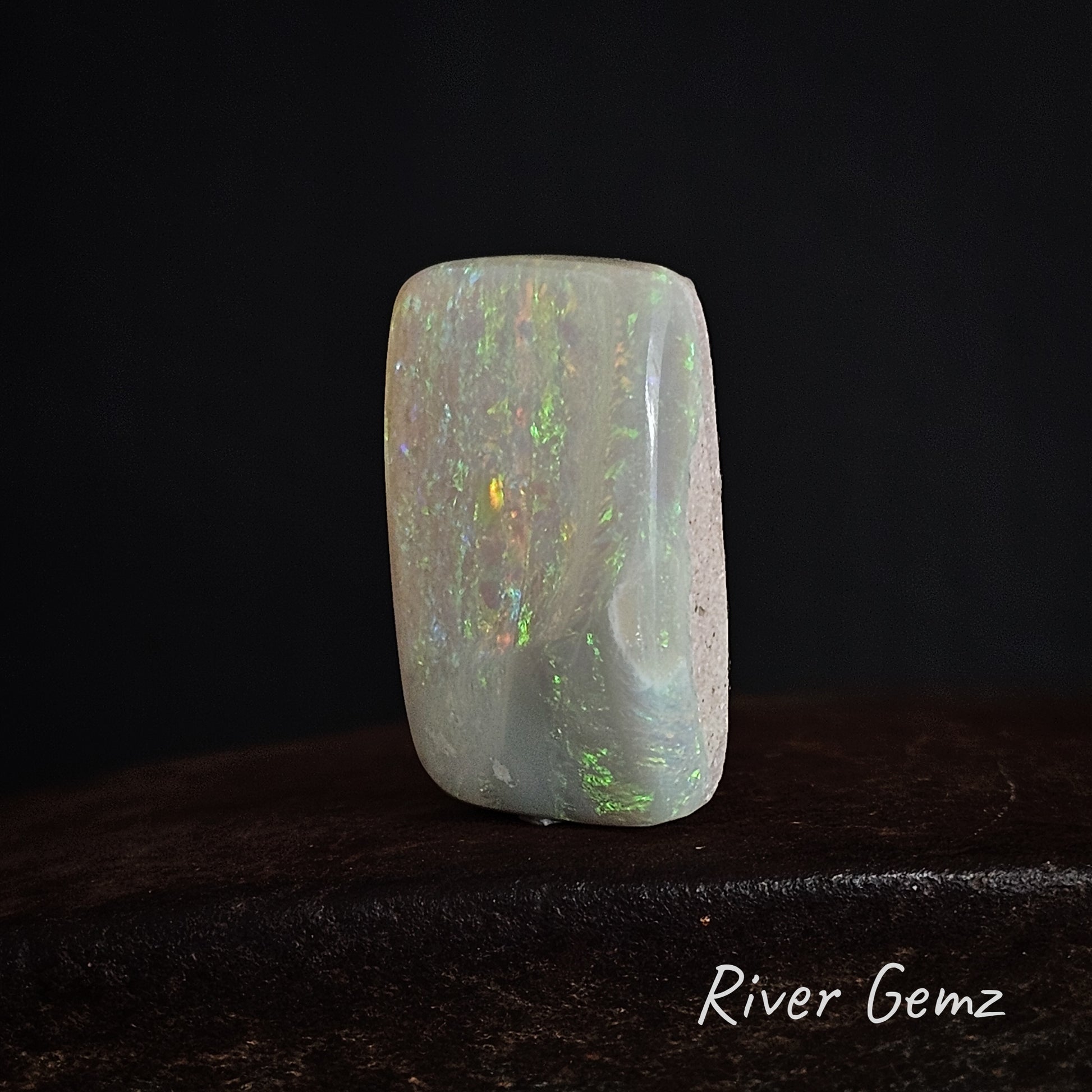 White unset opal stone with play of colours on a dark background, branded 'River Gemz'.