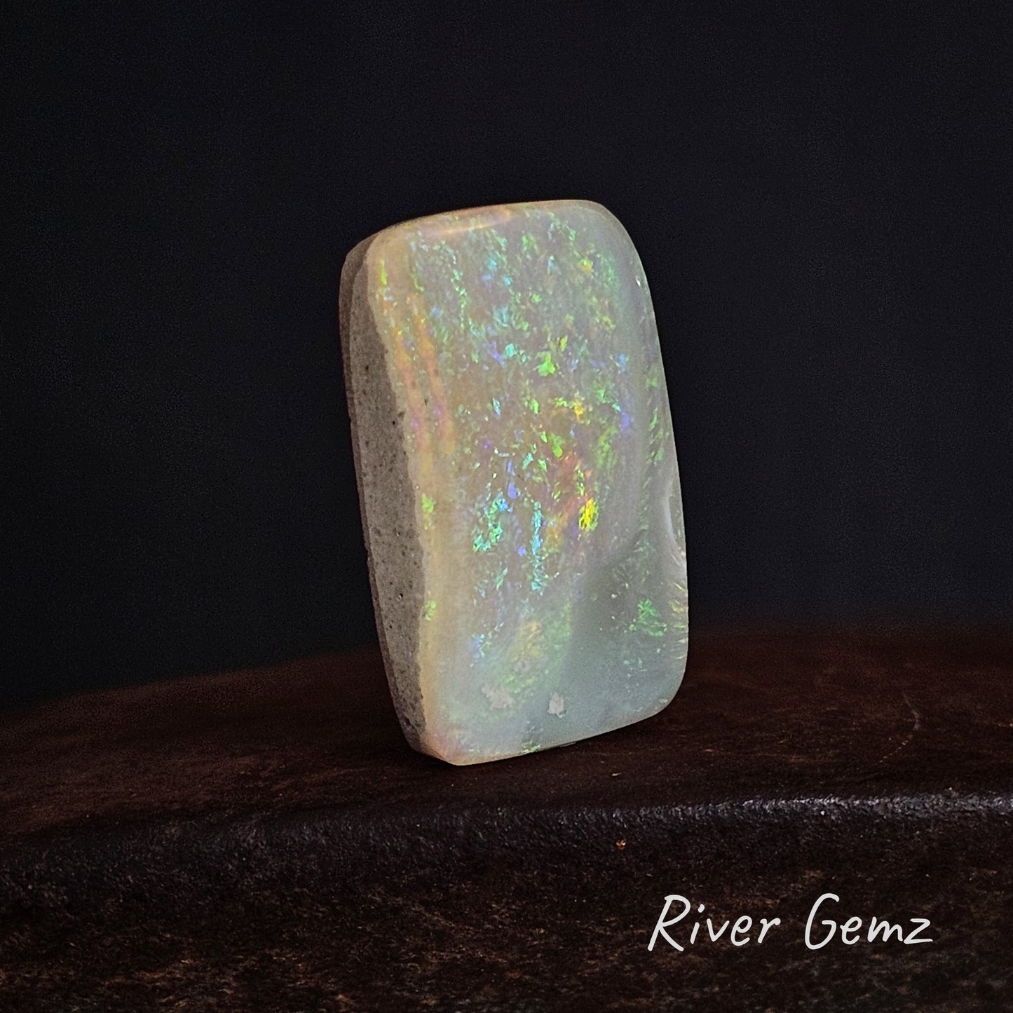 Rectangular light opal stone with multicoloured play of light on a dark background, branded 'River Gemz'.
