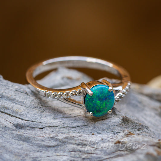 Ocean blue-green coloured circular black opal held securely with 4 claws evenly distributed around the circumference. Ring is resting on light-grey toned drift wood.