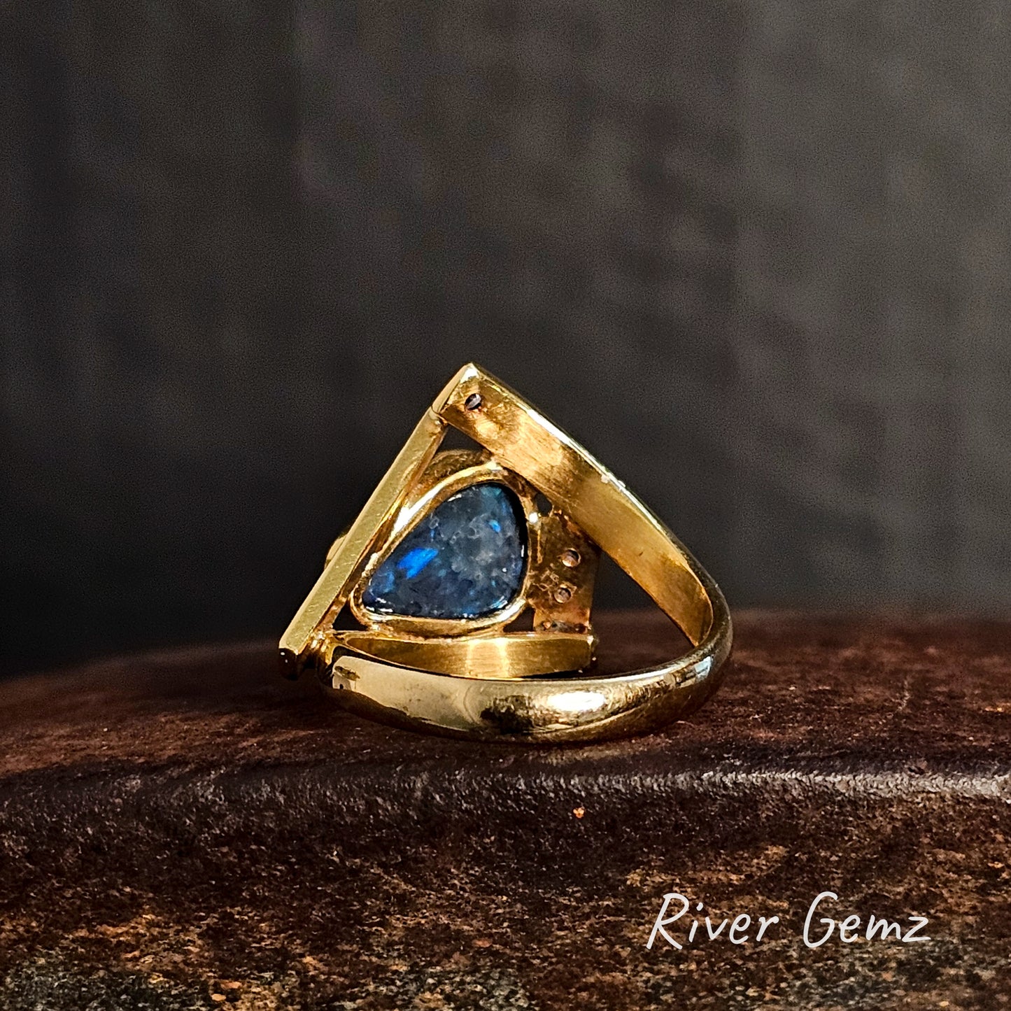 Back of the gold ring shows the dark base tone of the included black crystal opal on a cast iron surface, branded 'River Gemz'.