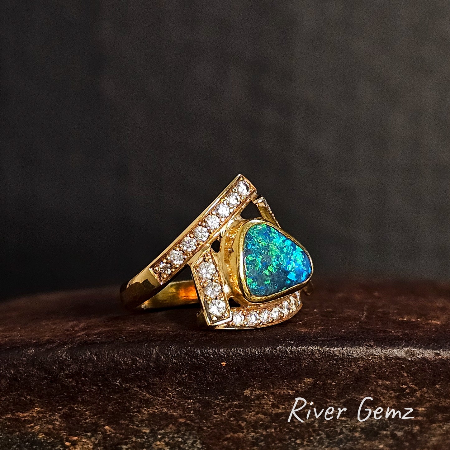 Gold ring with a blue and green black opal and diamonds on a dark surface, branded 'River Gemz'.