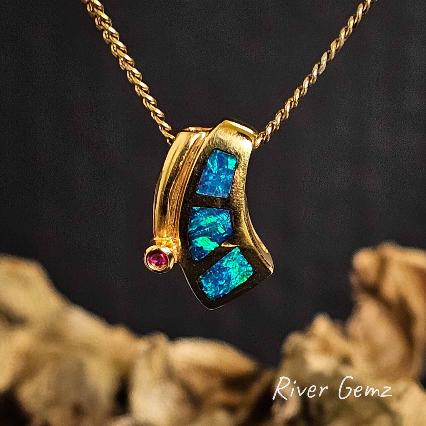 Gold pendant with blue/green black crystal opal and a ruby on a dark background, featuring 'River Gemz' branding.