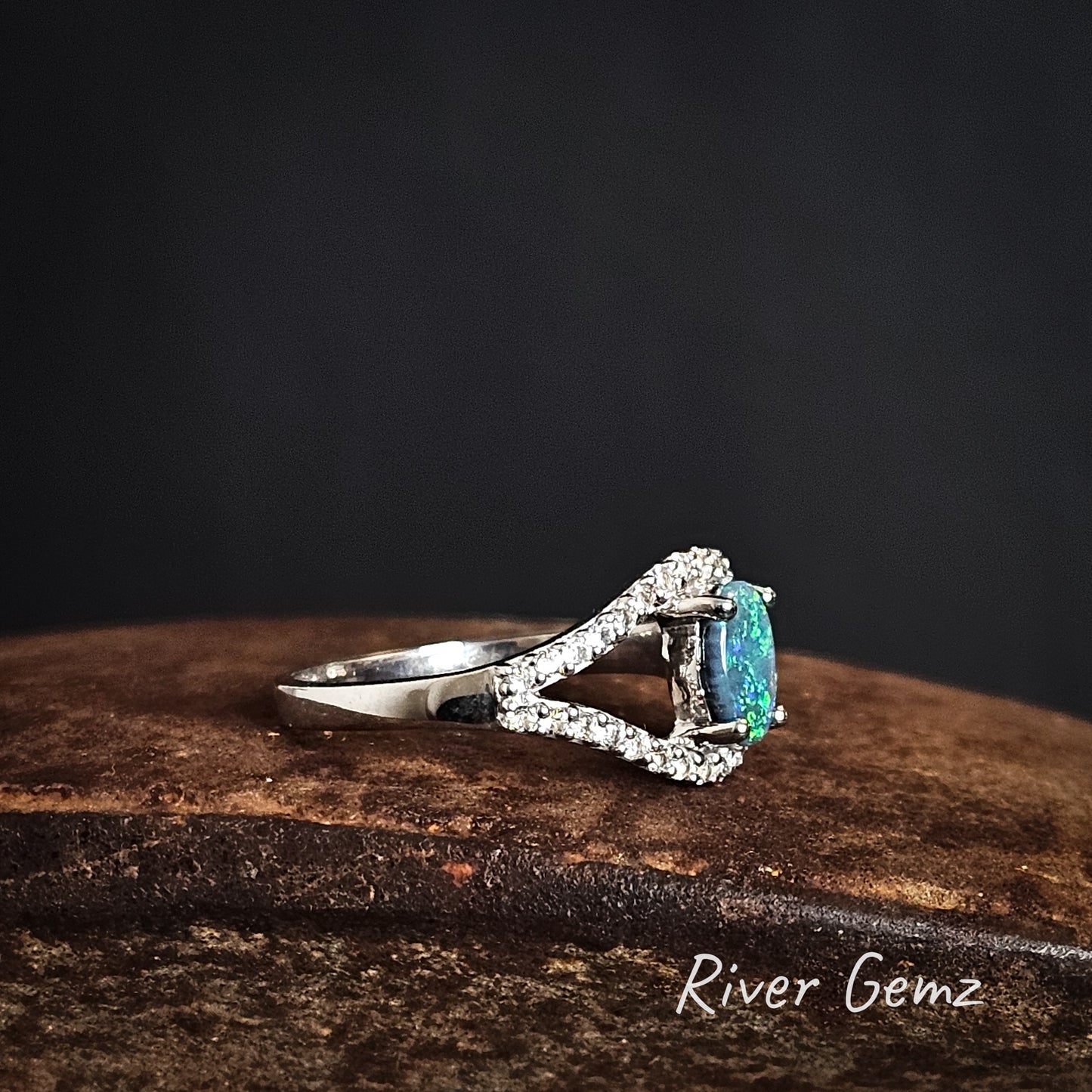 Side view of silver ring with a green black crystal opal and white topaz stones on a dark background, branded 'River Gemz'.