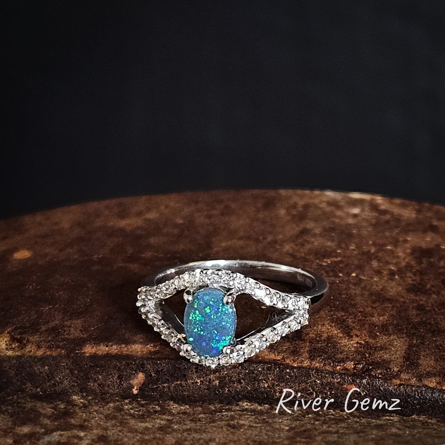 Silver ring with a blue and green oval shaped dark opal with two channels of topaz stones on a dark background, branded 'River Gemz'.