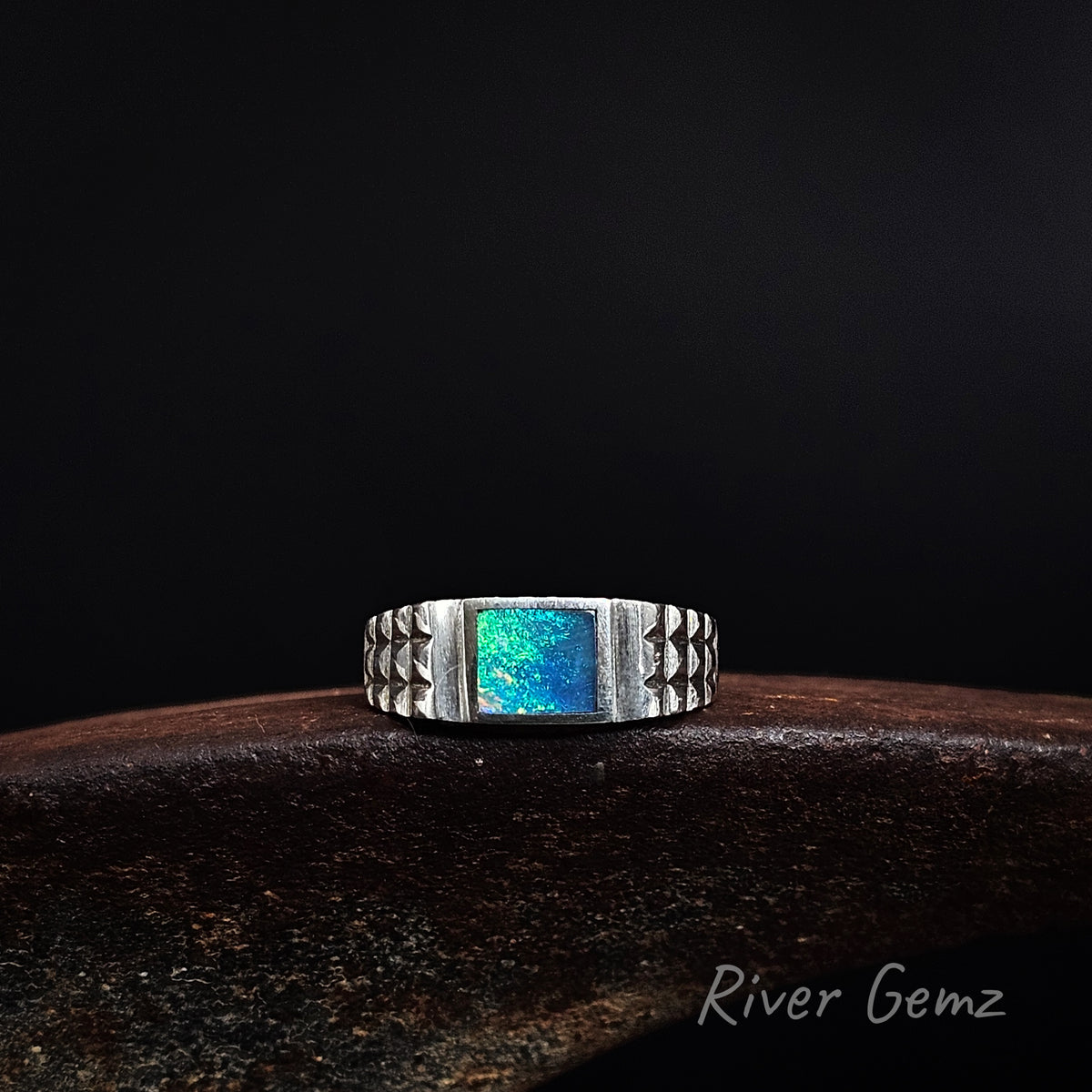 Blue and green dark opal inlayed in a men's sterling silver ring on a weathered shoe anvil and black background with 'River Gemz' watermark.