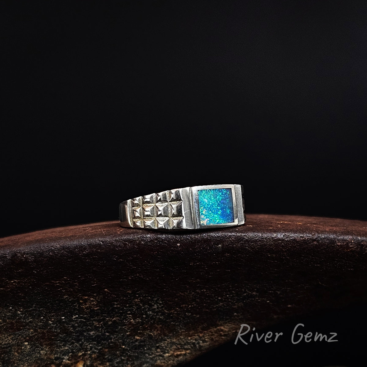 Slightly side on view of blue and green dark opal inlayed in a men's sterling silver ring on a weathered shoe anvil and black background with 'River Gemz' watermark.