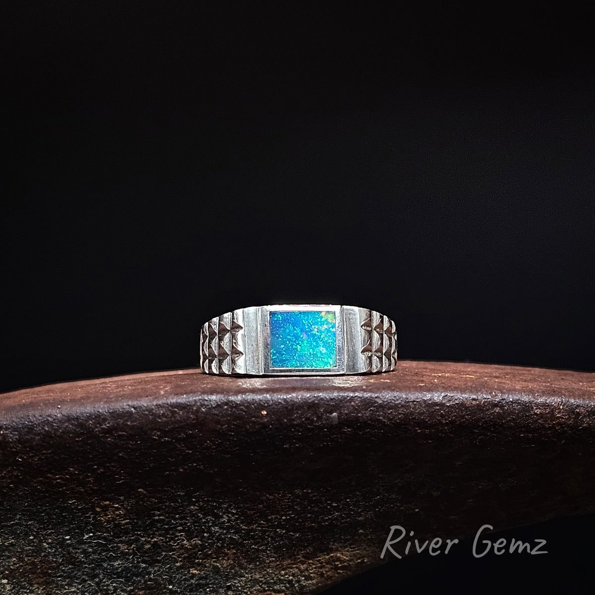Blue and green dark opal inlayed in a men's sterling silver ring on a weathered shoe anvil and black background with 'River Gemz' watermark.