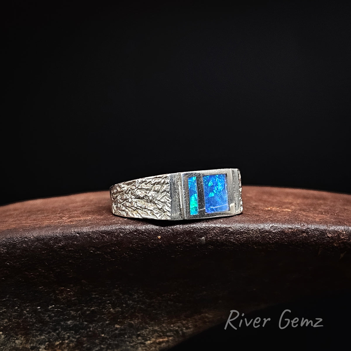A silver ring with a textured band with two vibrant blue opal gemstones, resting on a dark wooden surface against a black background. The brand name 'River Gemz' is visible in the bottom right corner.
