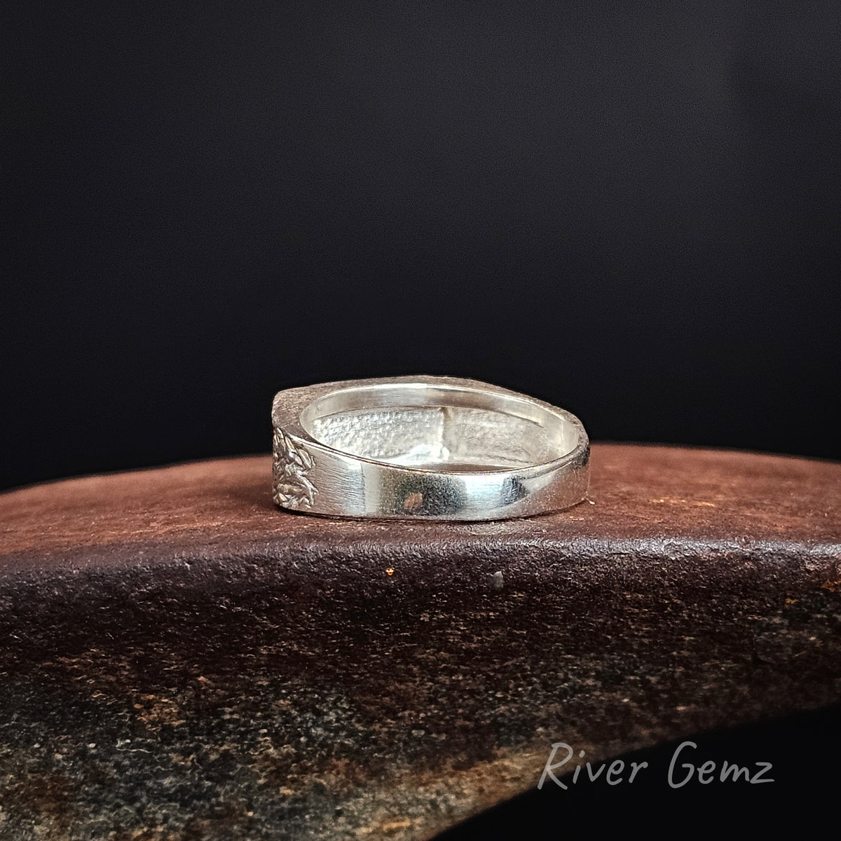 Rear view of a sterling silver showing the full silver backing behind the opal, resting on a rustic, dark brown anvil against a solid black background. The 'River Gemz' brand name is visible as a watermark in the bottom right corner.