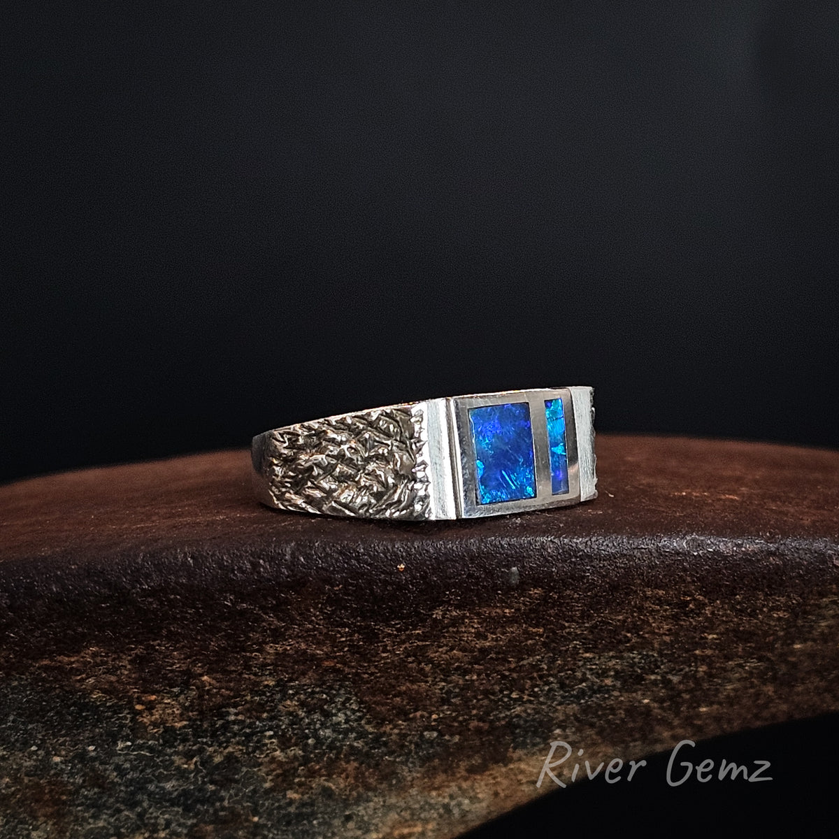 A silver ring with a textured band and a rectangular inlay featuring vibrant blue opal, resting on a dark wooden surface against a black background. The brand name 'River Gemz' is visible in the bottom right corner.