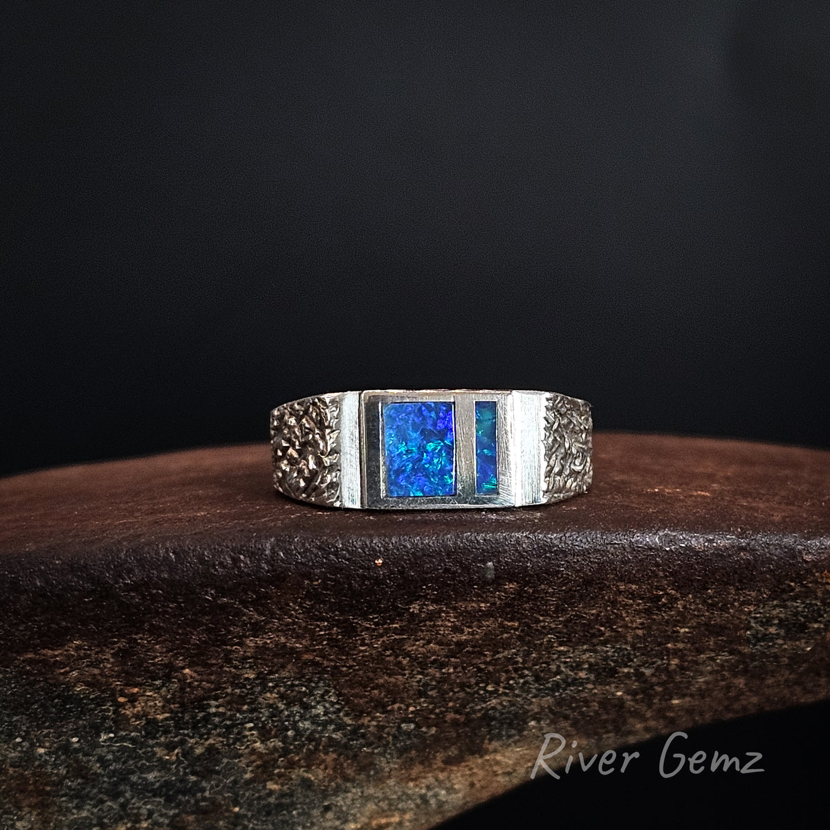 A silver-toned ring with two rectangular shaped opals inlaid, displayed on a dark textured surface against a black background. The brand name 'River Gemz' is visible in the bottom right corner.